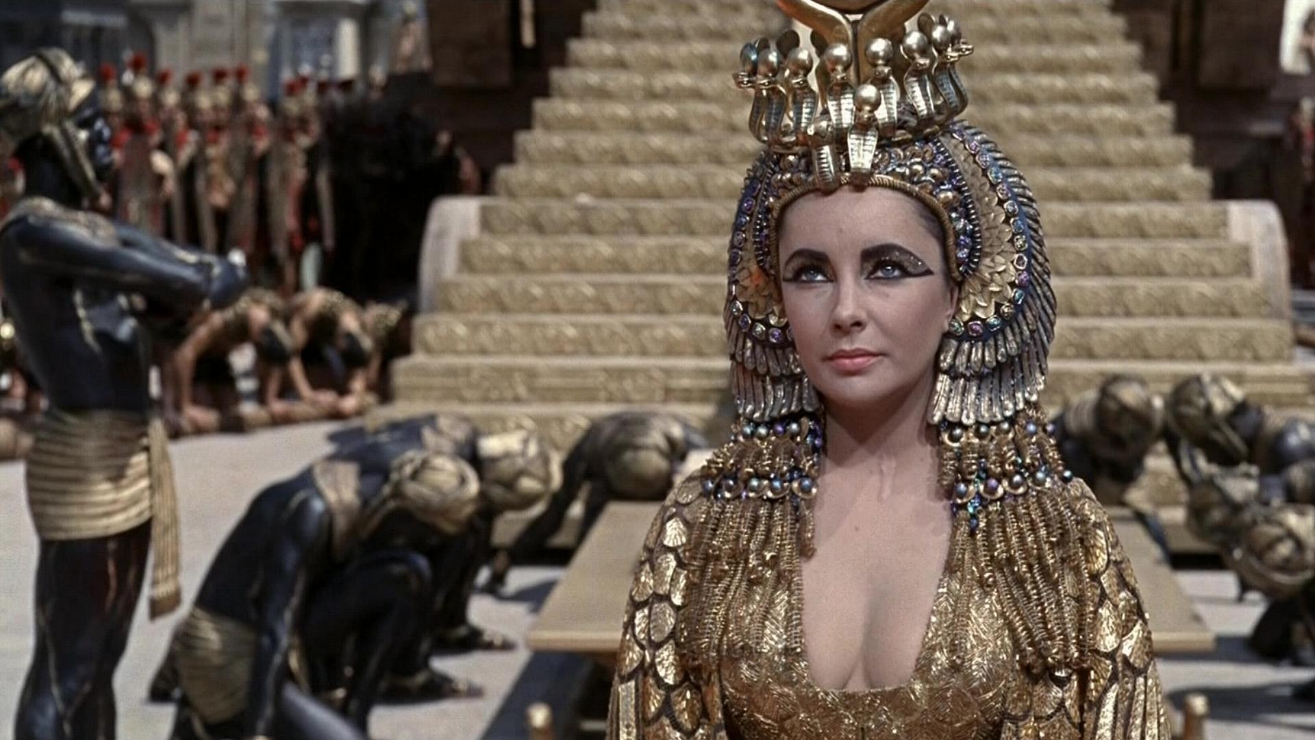 A woman in an elaborate ancient Egyptian costume stands in front of grand steps with kneeling figures.